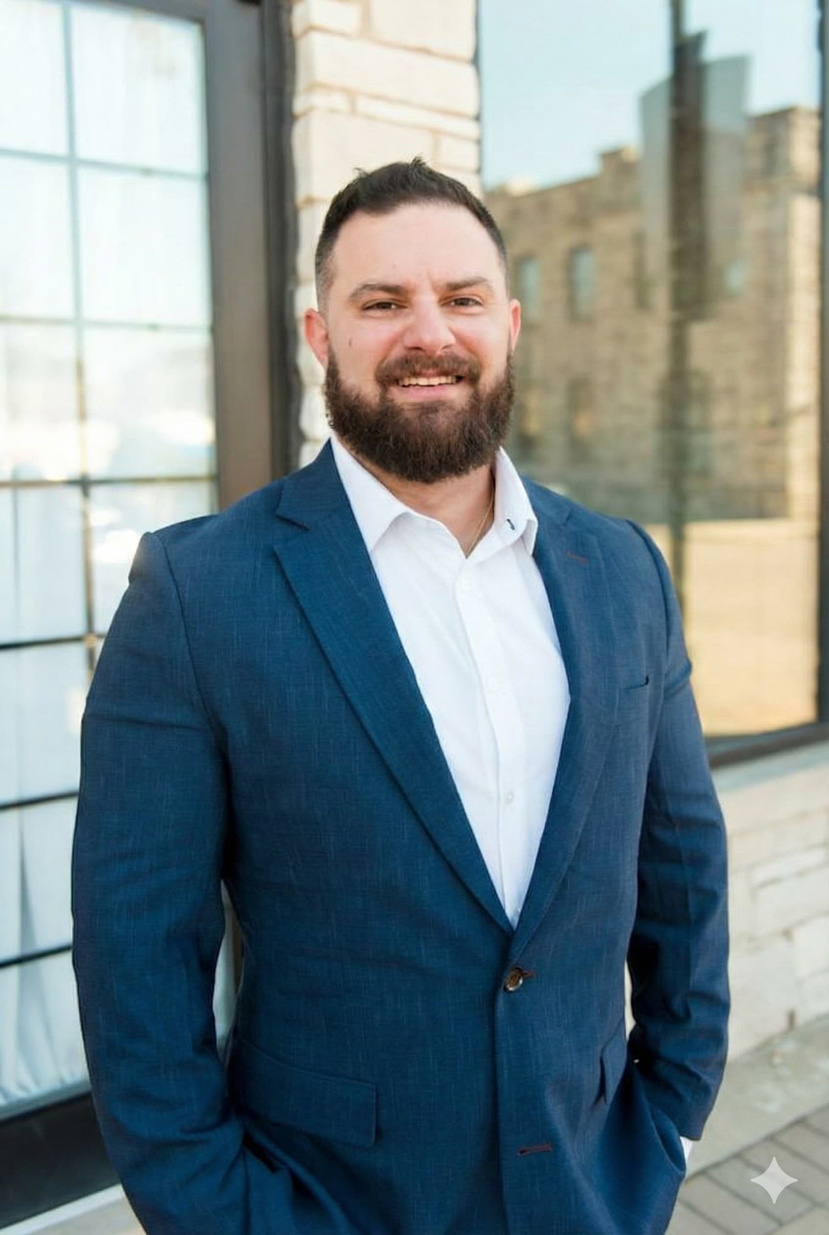 Smiling man with dark beard in blue suit and white shirt, standing outdoors.